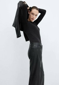 Top nero a maniche lunghe abbinato a pantaloni in pelle nera. La modella tiene in mano una giacca in pelle nera, evidenziando una silhouette aderente e una texture liscia.