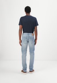 T-shirt à manches courtes marine porté avec un jean bleu clair, avec une patch en cuir sur la ceinture arrière. Des chaussures brunes complètent la tenue.