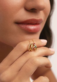 Gioielli in oro con due anelli; uno intrecciato e l'altro con gemme colorate. Primo piano di una mano che tiene gli anelli vicino alle labbra.