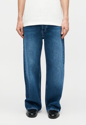 Osoba oblečená v voľných modrých rifliach v kombinácii s čiernymi koženými topánkami, stojaca na jednoduchom bielom pozadí.