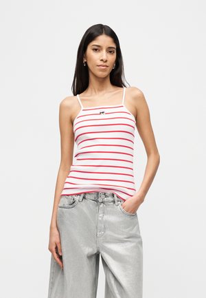 Donna con canottiera a spalline bianche e rosse a righe con una piccola grafica di un volto, abbinata a pantaloni larghi grigio chiaro, in piedi con una mano in tasca.