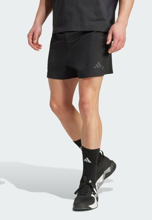 Persoon draagt zwarte sportshorts, zwarte enkelsokken en zwart-witte sportschoenen, staand met gekruiste benen.