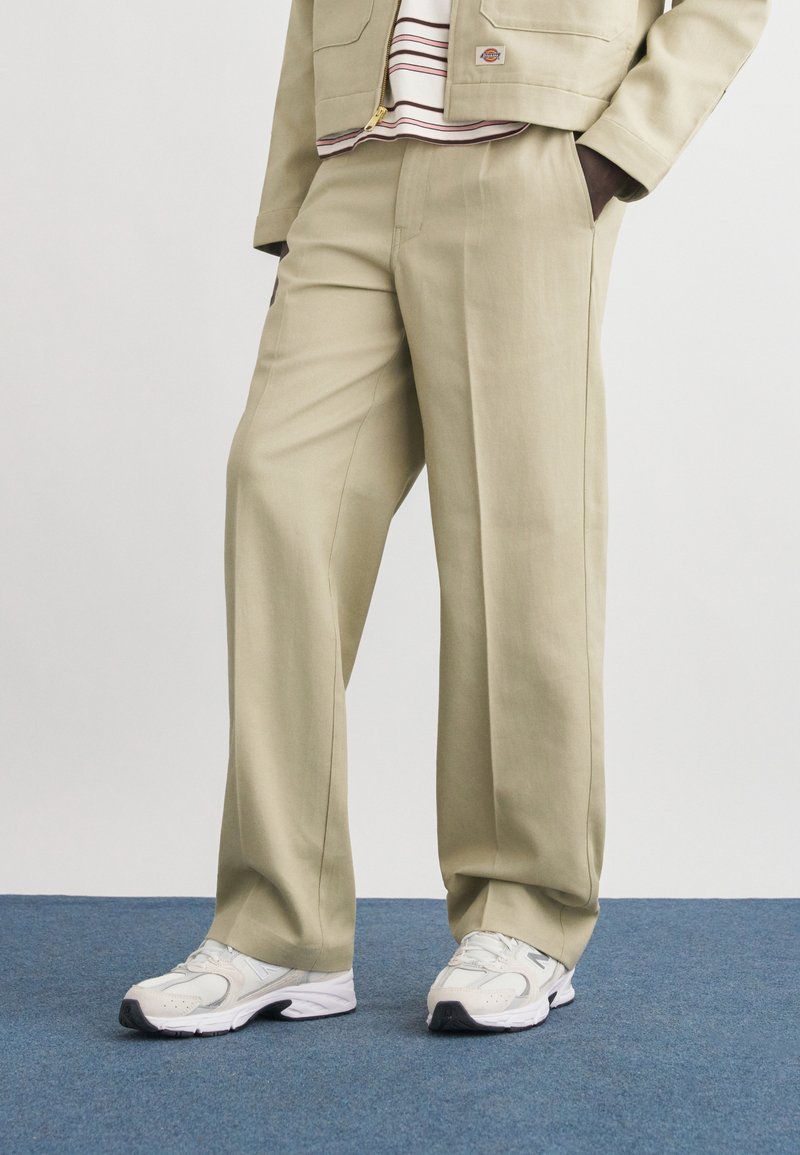 Κάτω μέρος σώματος με μπεζ φαρδιά παντελόνια, λευκά αθλητικά παπούτσια και μπεζ σακάκι πάνω από ριγέ πουκάμισο, στέκεται σε μπλε χαλί.