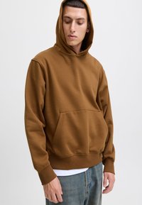 Brązowa bluza z dużą przednią kieszenią, z kapturem na sznurki i ściągaczami na rękawach, noszona na białej koszuli i w połączeniu z niebieskimi dżinsami.