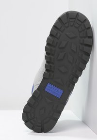 Semelle de chaussure Merrell en caoutchouc noir avec un motif de bande de roulement robuste et une étiquette bleue "Air Cushion" ; présente une partie supérieure en suède gris.