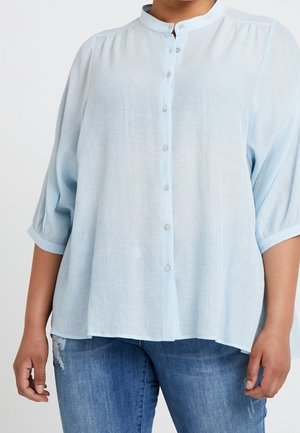 Blusa azul claro abotonada con cuello mandarín y mangas tres cuartos, confeccionada en tela ligera, presenta un ajuste relajado y una textura sutil.
