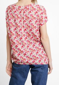 Blusa floral en rosa, rojo y amarillo con mangas cortas, escote redondo y detalle de pliegue en la parte posterior. Combinada con jeans de mezclilla azul.