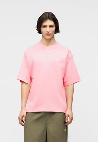 Νεαρό άτομο με σκούρα μαλλιά μέχρι τους ώμους, φορώντας ένα φαρδύ ροζ T-shirt Adidas και χακί σορτς, στέκεται μπροστά σε ένα απλό φόντο.