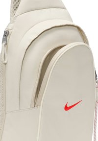 Beige Nike ryggsäck med en mjuk tygkonstruktion, dragkedjeförsedd framficka och en framträdande röd logotyp. Inkluderar vadderade remmar för komfort.