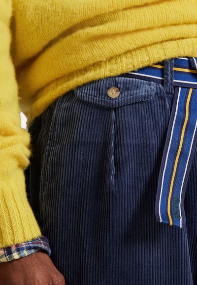 Marineblaue Cordhosen mit Vorderknopf, akzentuiertem Gürtel mit blau-gelben Streifen und strukturierter Oberfläche. Über der Hosen ist ein gelber Strickpullover sichtbar.