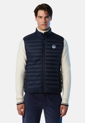 Uomo che indossa un gilet imbottito blu navy sopra un maglione a collo alto crema e pantaloni blu scuro, in piedi su uno sfondo bianco semplice.