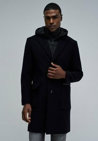 Abrigo largo de lana negra con solapas muestradas, dos bolsillos frontales y cierre de botones. Lleva encima un hoodie negro con cremallera. Textura suave.