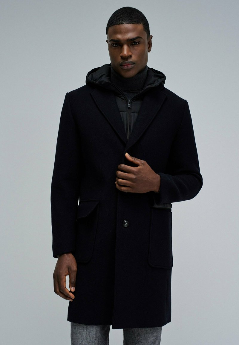 Abrigo largo de lana negra con solapas muestradas, dos bolsillos frontales y cierre de botones. Lleva encima un hoodie negro con cremallera. Textura suave.