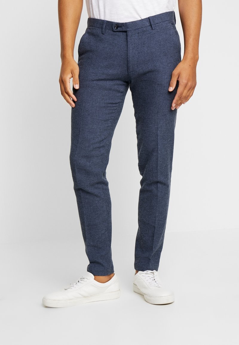 Tailored navy trousers in textured fabric, featuring a slim fit, belt loops, front pockets, and a button closure. Paired with white sneakers.