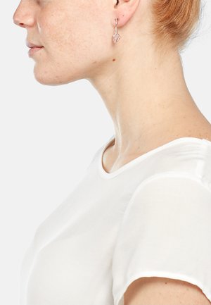 Boucles d'oreilles pendantes en or rose avec des motifs complexes, arborant une forme courbée et une texture lisse, portées par un modèle à la peau claire et aux cheveux roux.
