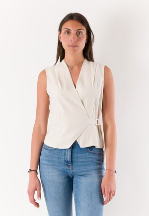 Donna con lunghi capelli scuri indossa un top bianco senza maniche con fibbia e jeans blu, in piedi davanti a uno sfondo chiaro e uniforme.