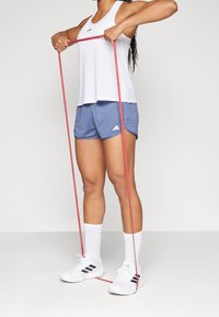 Mujer con una camiseta sin mangas blanca y pantalones cortos azules haciendo ejercicio con una banda de resistencia roja, de pie sobre un suelo y fondo blanco liso.