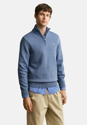 Jeune homme portant un pull bleu à demi-fermeture éclair par-dessus une chemise rayée bleue et un pantalon beige, debout devant un fond blanc.