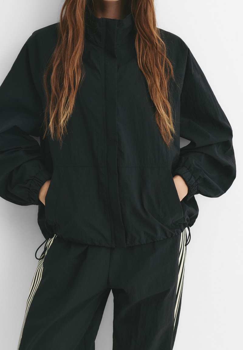Femme portant une veste légère noire avec des poignets élastiques et un pantalon noir assorti avec des bandes blanches sur les côtés, mains dans les poches, sur fond blanc.
