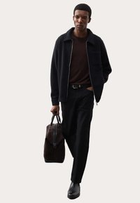Giacca nera con zip e colletto, maglione marrone sottostante, pantaloni scuri e scarpe nere. Porta una borsa a tracolla marrone con dettagli neri.