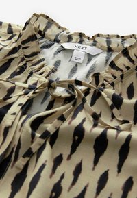 Lettet beige bluse med sorte og brune abstrakte mønstre, flæset halsudskæring og bundet front. Etiketten siger "NEXT," lavet i Indien.
