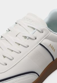Λευκό χαμηλό sneaker ARKK με λευκά κορδόνια, διάτρητη πλευρική λεπτομέρεια, ναυτικό μπλε περιθώριο και καφέ λαστιχένια σόλα.