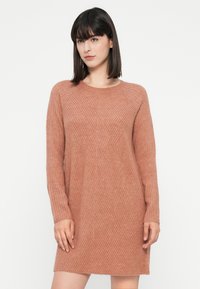Žena ve svetle růžových šatech s dlouhými rukávy a žebrovanou pleteninou stojící na bílém pozadí.