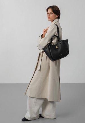 Vrouw in een beige trenchcoat en brede witte broek draagt een grote zwarte handtas over haar schouder, staand tegen een effen grijze achtergrond.