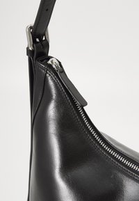 Sac en cuir noir avec une finition brillante, doté d'une fermeture éclair et de quincaillerie en argent. Forme incurvée avec une bandoulière ajustable. Design minimaliste.