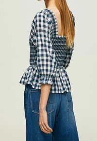 Blusa di cotone a quadretti con maniche a puff, busto aderente e orlo a peplo svasato; combinazione di colori blu navy e bianco con dettagli arricciati.