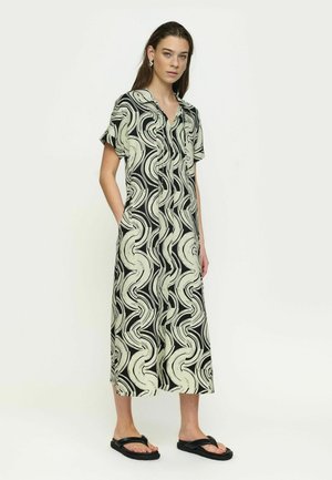 Žena s dlhými vlasmi, ktorá nosí midi šaty s krátkymi rukávmi v čiernej a béžovej farbe so vzorom víru a čierne sandále, stojaca pred hladkým pozadím.