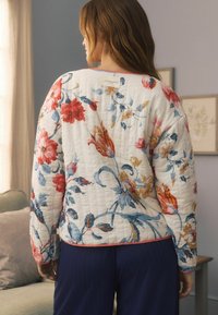 Giacca patchwork floreale in tessuto crema, con motivi di fiori rossi e blu, un colletto rotondo e una rifinitura rosa lungo le maniche e l'orlo.