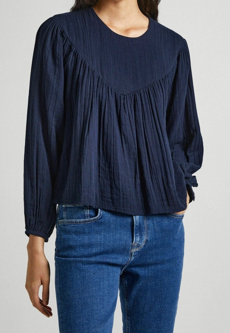 Femme portant un chemisier à manches longues bleu marine avec des détails plissés et un jean denim bleu, une main dans la poche.