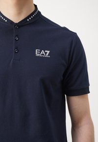 Navy polo shirt made of textured fabric featuring a buttoned placket, short sleeves, and a contrasting collar with patterned accents.