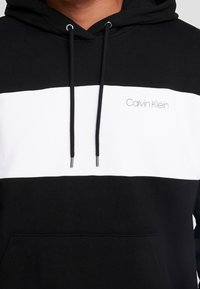Felpa con cappuccio nera e bianca con cordino, tasca frontale e logo "Calvin Klein" sul petto, realizzata in tessuto morbido.