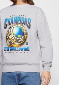 Felpa grigia con una grande grafica che presenta la parola "CHAMPIONS" sopra un globo e un anello del campione. Gli accenti includono un testo blu e oro in grassetto.