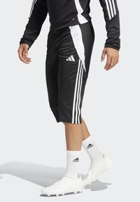 Svarte sportsshorts med hvite side-striper, laget av glatt stoff. Kombinert med hvite fotballsko og stripete hvite sokker.