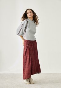 Suéter gris claro acanalado con detalles en burdeos, combinado con una falda larga de burdeos con estampado y deportivas beige, sobre un fondo neutro.