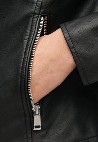 Χέρι μερικώς εισαγόμενο στην ανοιχτή τσέπη μαύρου υφής δερμάτινου μπουφάν με ορατό ασημένιο φερμουάρ.