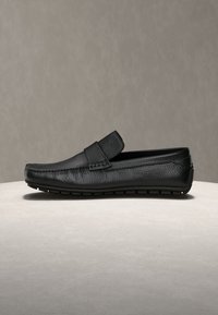 Mocassino nero in pelle senza lacci con tomaia testurizzata e suola in gomma, esposto su una superficie rotonda chiara contro uno sfondo neutro.