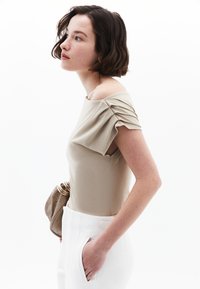 Beige off-shoulder top med plisserede korte ærmer, parret med højhalsede hvide bukser med sidelommer. Brun clutch taske.