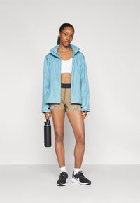 Donna con giacca blu, reggiseno sportivo bianco, pantaloni corti a righe, sneakers nere, che tiene una bottiglia d'acqua nera, in piedi su uno sfondo neutro.