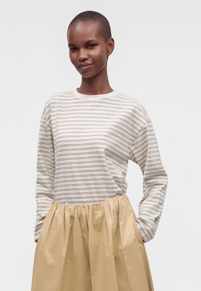 Femme aux cheveux courts portant une chemise blanche à manches longues avec de fines rayures noires et une jupe beige avec une taille froncée, debout devant un fond uni.
