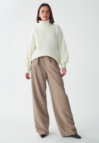 Maglione a collo alto di colore crema con maniche ampie abbinato a pantaloni larghi beige a vita alta e décolleté neri a punta.