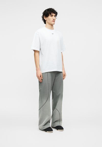 Νέος άνδρας με κατσαρά μαλλιά, που φοράει λευκό φαρδύ T-shirt και άνετο γκρι παντελόνι με μαύρες ραφές, στέκεται μπροστά σε λευκό φόντο.