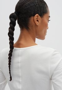 Weißes Langarmoberteil mit einem rundem Halsausschnitt, aus einem glatten, strukturierten Stoff. Die Haare sind in einem gedrehten Zopf frisiert.