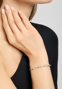 Bracelet en argent avec des maillons ronds et ovales, ornée de pierres précieuses transparentes. Texture lisse, porté sur un poignet avec du vernis à ongles neutre.