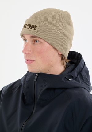 Khaki neulepipo, jossa on ribbikuviointi ja ommeltu "SLOPE" -merkki. Yhdistettynä tummansiniseen vetoketjulliseen takkiin, jossa on korkea kaulus ja tuuletusaukot.