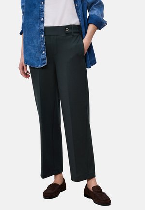 Osoba ubrana w czarne spodnie o prostych nogawkach z pętlą na pasek zapinaną na guziki, niebieską denimową koszulę, biały top oraz ciemnobrązowe mokasyny. Jedna ręka w kieszeni.