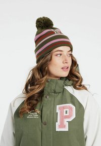 Zelená a biela parka s ružovým znakom "P", vysokým golierom a gombíkovým zapínaním; hnedá a zelená pruhovaná pletená čiapka s pompomom.
